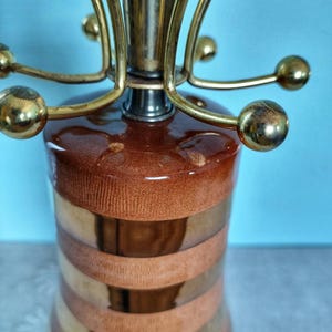 K&ouml;nnte beinhalten: Eine Vintage-Tischlampe in Braun- und Goldt&ouml;nen. Die Lampe hat einen zylindrischen Sockel mit horizontalen B&auml;ndern in Braun und Gold. Die Oberseite hat eine Messinghalterung mit gebogenen Armen und kugelf&ouml;rmigen Akzenten. Dekorative Beleuchtung.