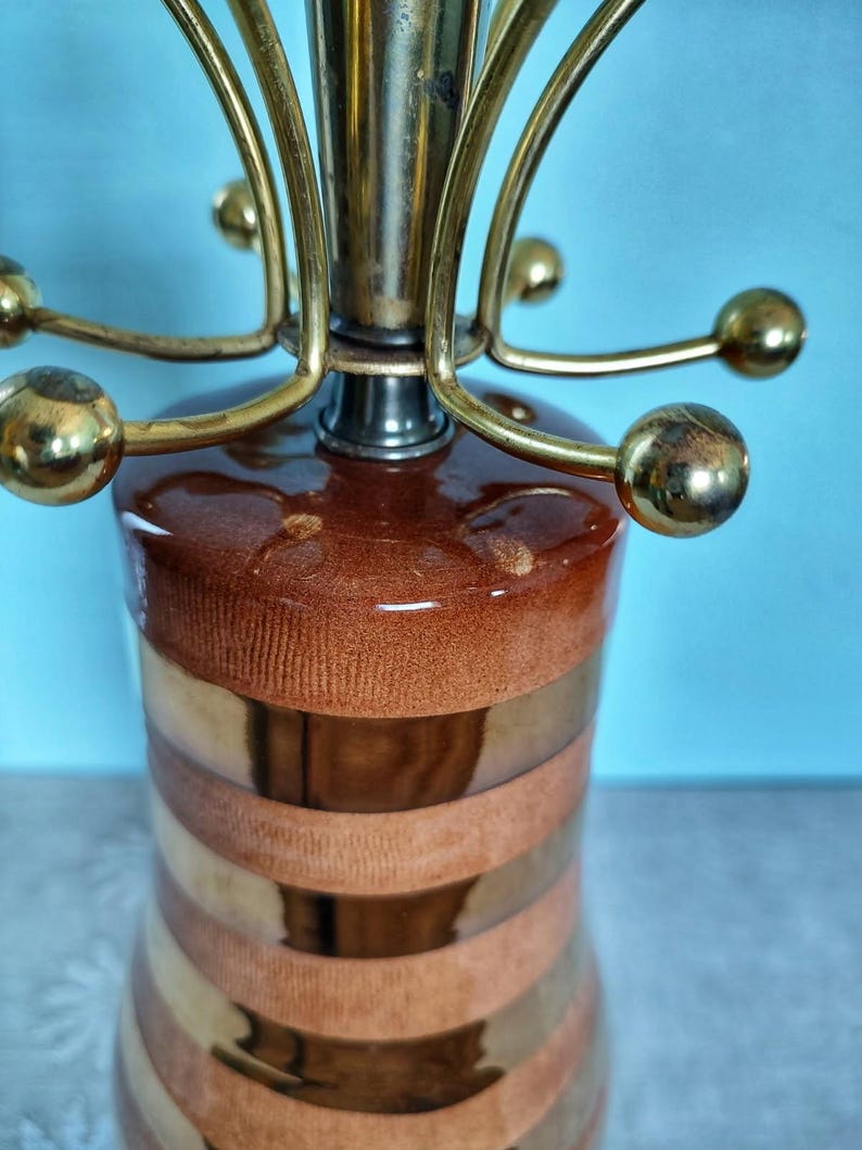 K&ouml;nnte beinhalten: Eine Vintage-Tischlampe in Braun- und Goldt&ouml;nen. Die Lampe hat einen zylindrischen Sockel mit horizontalen B&auml;ndern in Braun und Gold. Die Oberseite hat eine Messinghalterung mit gebogenen Armen und kugelf&ouml;rmigen Akzenten. Dekorative Beleuchtung.