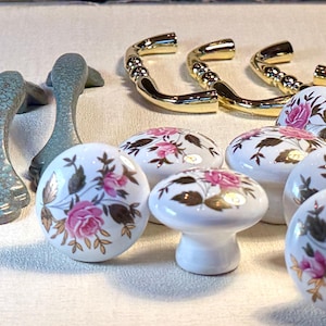 May include: A collection of cabinet hardware, including gray handles, gold-toned pulls, and white ceramic knobs. The knobs feature a pink rose and gold leaf design. The hardware is suitable for furniture restoration or new builds.