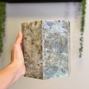 May include: A large, hexagonal labradorite crystal. The stone has a mottled appearance with shades of gray, beige, and blue. The crystal is held in a hand, showcasing its size and natural beauty. The polished surfaces reflect light.