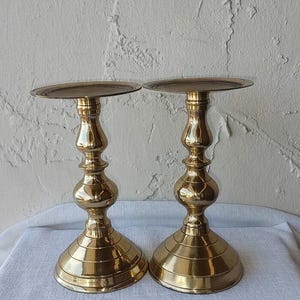May include: A pair of polished brass candle holders featuring a wide circular base, a slender stem with a rounded central bulbous section, and a flat circular top plate designed to support a candle. The metal surface has a reflective gold finish.