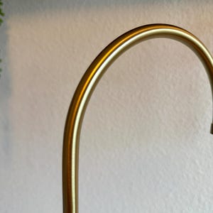 May include: A curved, gold-colored metal object, possibly a part of a lamp or decorative element. The smooth, polished surface reflects light, creating highlights. The background is a textured, off-white wall.