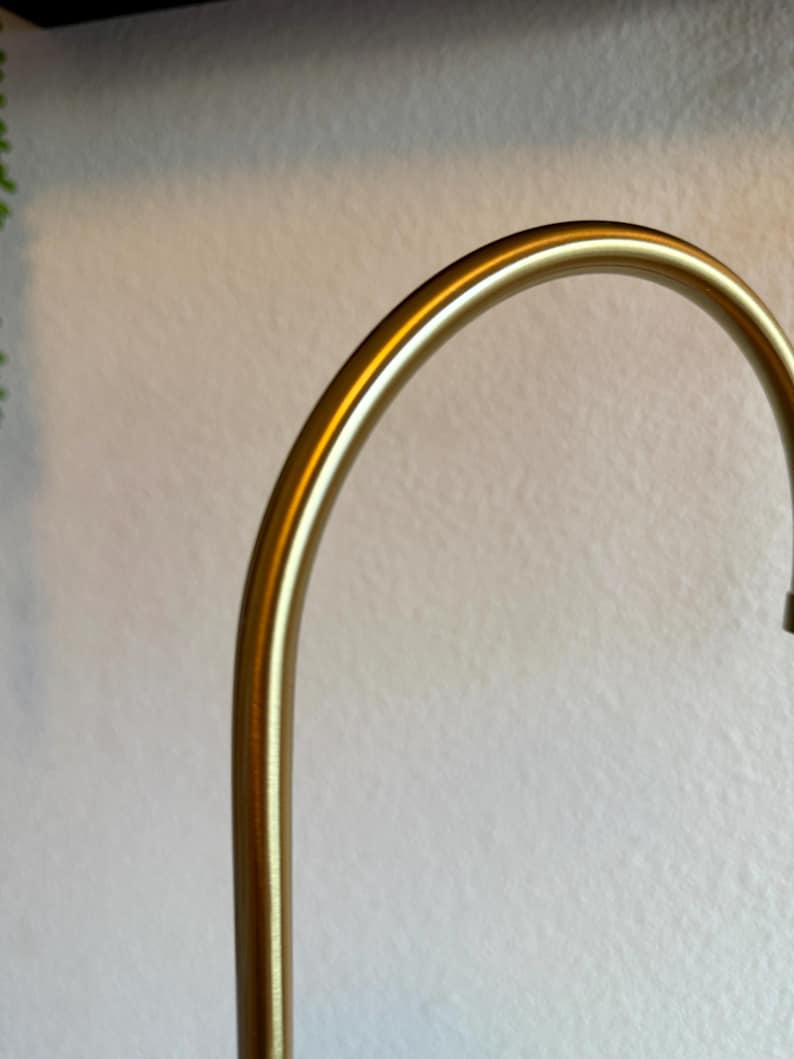 May include: A curved, gold-colored metal object, possibly a part of a lamp or decorative element. The smooth, polished surface reflects light, creating highlights. The background is a textured, off-white wall.