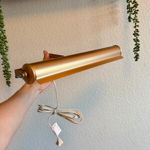 May include: A gold-colored picture light with a cylindrical body and a rectangular shade. The light is held by a hand, and a white cord with a plug extends from the base. The light is designed to be mounted on a wall.