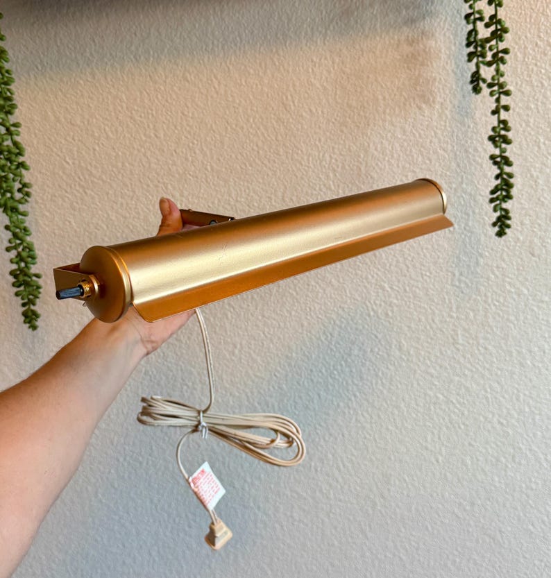 May include: A gold-colored picture light with a cylindrical body and a rectangular shade. The light is held by a hand, and a white cord with a plug extends from the base. The light is designed to be mounted on a wall.