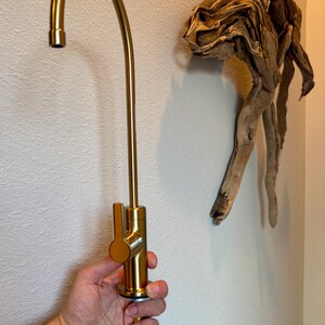 May include: A gold-colored kitchen faucet with a curved spout and lever handle is held against a white wall. A piece of driftwood art is mounted on the wall to the right of the faucet.