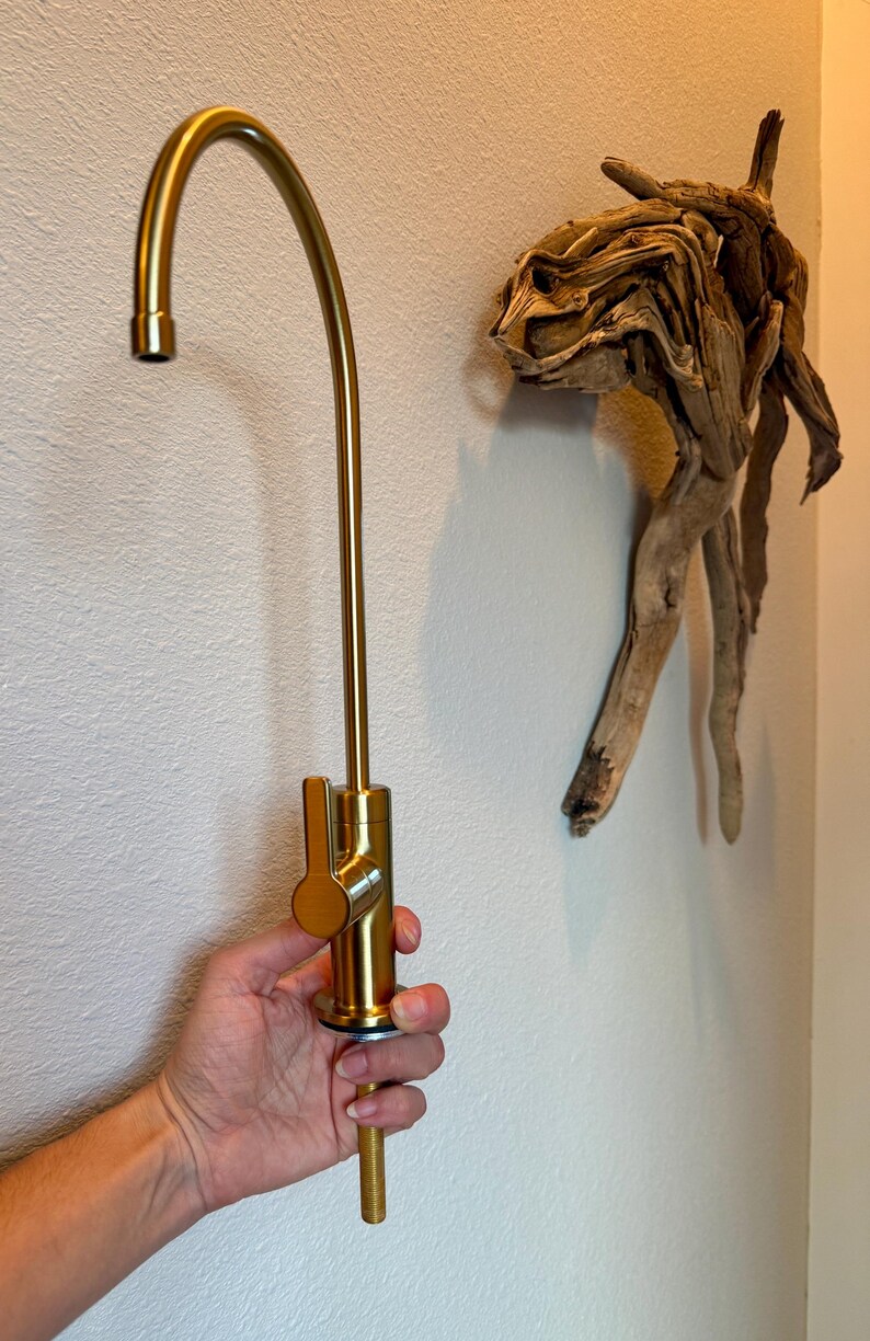 May include: A gold-colored kitchen faucet with a curved spout and lever handle is held against a white wall. A piece of driftwood art is mounted on the wall to the right of the faucet.