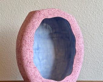 May include: A ceramic vase with an oval shape and a textured, speckled pink exterior. The interior of the vase features a smooth, light blue finish with a wavy, irregular opening that creates a deep, hollow cavity within the center of the object.