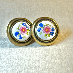 May include: Two vintage cabinet knobs with floral designs. Each knob features a white ceramic center with pink, blue, and green flowers, surrounded by a gold-toned metal frame. The knobs are round and likely used for furniture.