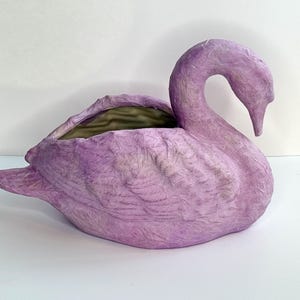 May include: A lavender-colored swan-shaped planter. The swan has a curved neck and a detailed feather texture. The interior of the planter is a darker green. The planter is made of a textured material and sits on a white surface.