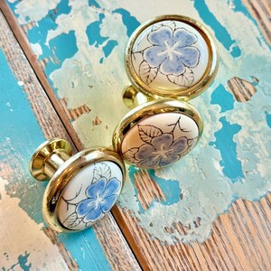 May include: Three round cabinet knobs featuring a white ceramic center with a hand-painted blue flower and black leaf outlines. Each knob is encased in a polished gold-tone metal rim and includes a matching gold-tone metal base for mounting.