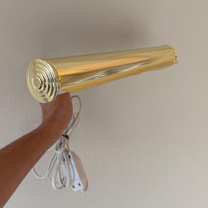 May include: A gold-colored, cylindrical wall-mounted light fixture. The light has a circular end cap with concentric rings and a white power cord with a switch. The fixture is designed to be mounted on a wall.