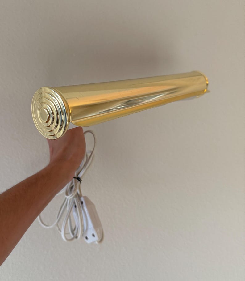 May include: A gold-colored, cylindrical wall-mounted light fixture. The light has a circular end cap with concentric rings and a white power cord with a switch. The fixture is designed to be mounted on a wall.