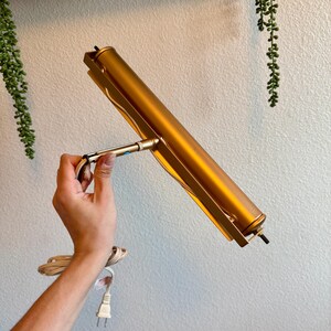 May include: A gold-colored picture light with a cylindrical body and a rectangular frame. The light is held by a hand, and a white electrical cord with a plug is visible.