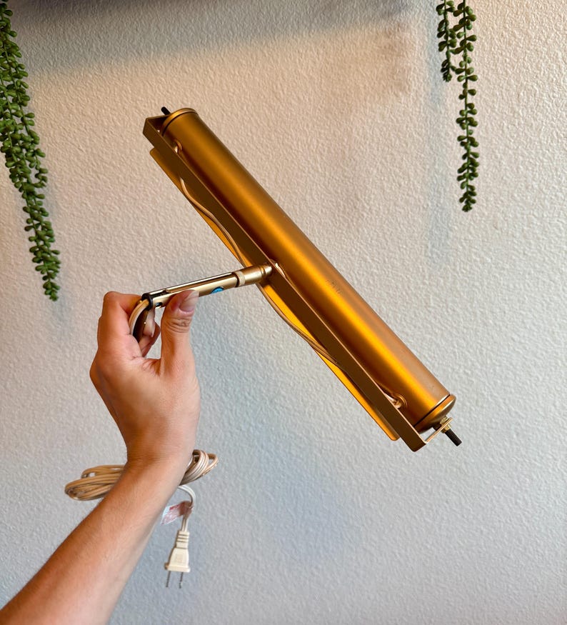 May include: A gold-colored picture light with a cylindrical body and a rectangular frame. The light is held by a hand, and a white electrical cord with a plug is visible.