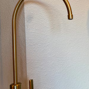 May include: A gold-colored kitchen faucet with a curved spout and a single lever handle. The faucet is made of metal and has a modern design. The faucet is mounted on a white wall.