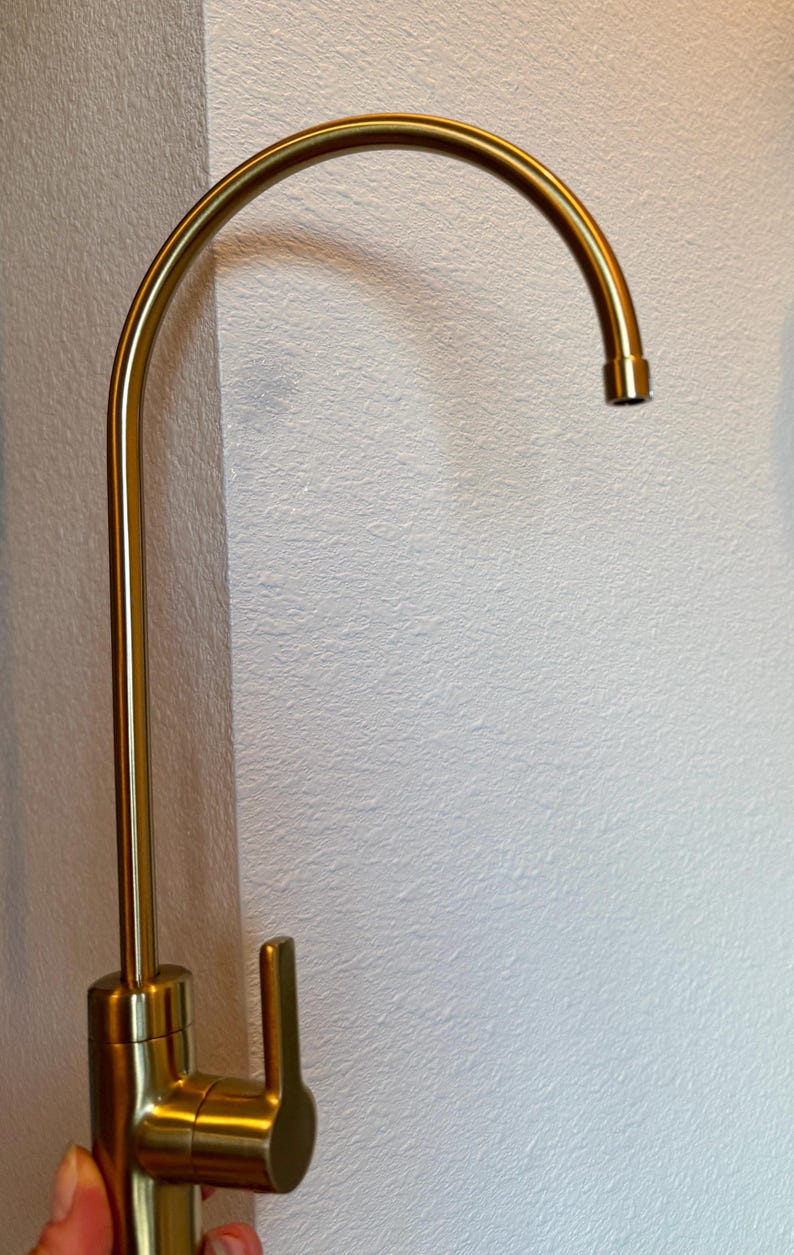 May include: A gold-colored kitchen faucet with a curved spout and a single lever handle. The faucet is made of metal and has a modern design. The faucet is mounted on a white wall.