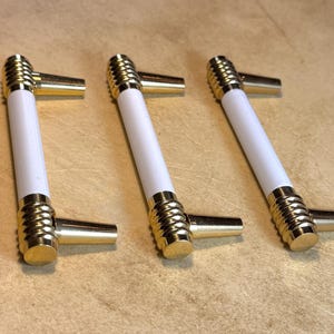 May include: Three cabinet pulls with a white center and gold-colored accents. The pulls have a cylindrical design with a ribbed texture on the ends and a rectangular handle. The hardware is designed for furniture and home decor.