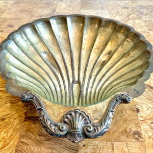 May include: A vintage, shell-shaped dish with a scalloped edge. The dish is made of a tarnished, gold-toned metal with a decorative, ornate handle. The interior has a ribbed design, and the dish is likely used for decorative purposes.