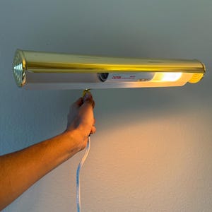 May include: A gold-colored picture light fixture with a white interior and a bright light. The fixture is mounted on a wall and is plugged in. The word "CAUTION" is visible on the fixture.