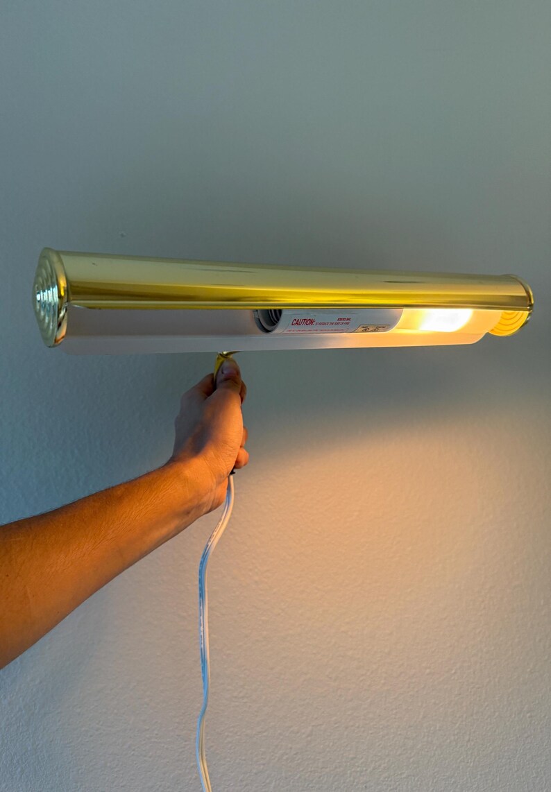 May include: A gold-colored picture light fixture with a white interior and a bright light. The fixture is mounted on a wall and is plugged in. The word "CAUTION" is visible on the fixture.