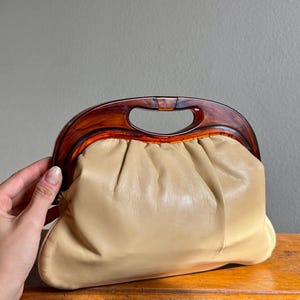 May include: A vintage beige leather clutch with a tortoiseshell handle. The bag has a gathered design and a curved, arched handle. The clutch is sitting on a wooden surface.