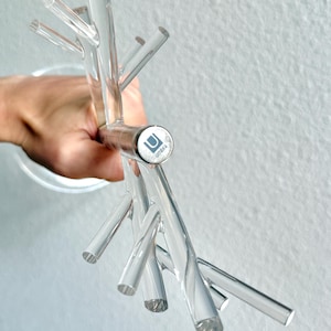 May include: Clear acrylic wall-mounted coat rack with multiple branches. The rack is held by a hand and features the Umbra logo on a silver-colored circular base. The design resembles a tree branch.