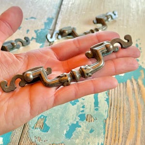 May include: A metallic drawer pull with a curved, ornate design featuring a dark, aged finish. The handle has a central grip section with decorative detailing and scrolled ends that extend outward, showcasing a weathered, antique-style appearance.