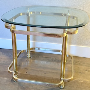 May include: A vintage side table with a clear glass top and a gold-toned metal frame. The table has a rectangular shape with rounded corners and a lower shelf. The legs are straight and connect to the top and bottom frames.