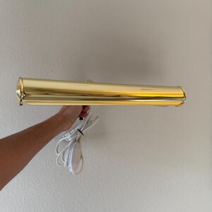 May include: A gold-colored, rectangular wall-mounted picture light. The light has a cylindrical shape with a decorative end and a white power cord. The light is held by a person's hand.