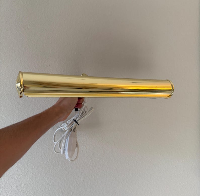 May include: A gold-colored, rectangular wall-mounted picture light. The light has a cylindrical shape with a decorative end and a white power cord. The light is held by a person's hand.
