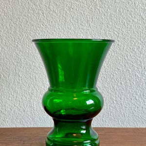 Könnte beinhalten: Eine leuchtend smaragdgrüne Glasvase mit einem breiten, ausgestellten Rand, der sich zu einem abgerundeten, bauchigen Mittelteil verjüngt und auf einem stabilen, kreisförmigen Sockel ruht. Das Glas ist glatt, durchscheinend und von einheitlicher grüner Farbe.