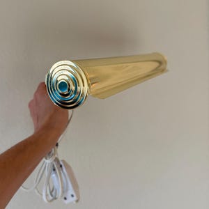 May include: A vintage brass wall-mounted light fixture. The fixture has a cylindrical body and a decorative end with concentric circles and a teal center. It includes a white cord and a plug.