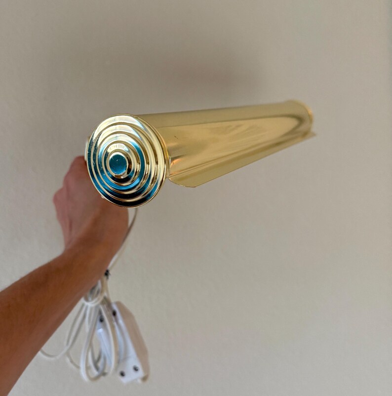 May include: A vintage brass wall-mounted light fixture. The fixture has a cylindrical body and a decorative end with concentric circles and a teal center. It includes a white cord and a plug.