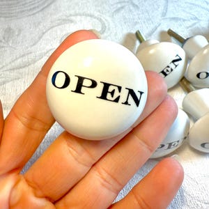 May include: White ceramic cabinet knobs with the word "OPEN" in black lettering. The knobs are round and have a brass screw. Other knobs with the letters "EN" and "O" are also visible.