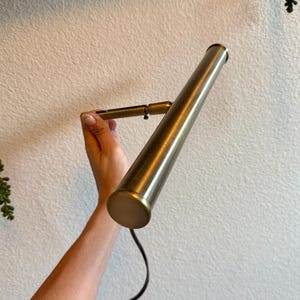May include: A brass-colored, adjustable wall-mounted lamp with a cylindrical shade. The lamp is held against a white wall, with a black cord extending from the base. The lamp's design is simple and modern, suitable for various interior styles.