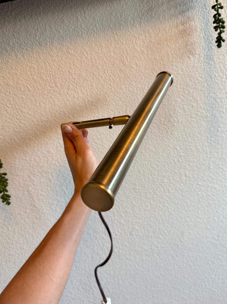 May include: A brass-colored, adjustable wall-mounted lamp with a cylindrical shade. The lamp is held against a white wall, with a black cord extending from the base. The lamp's design is simple and modern, suitable for various interior styles.