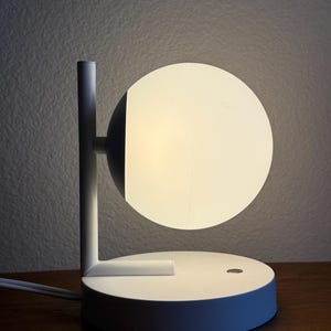 May include: A white table lamp featuring a circular, glowing glass globe shade attached to a vertical post. The lamp has a flat, round base with a small circular power button on the surface and a white power cord extending from the back of the base.