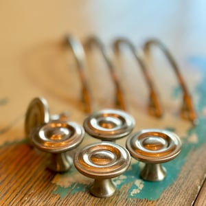 May include: A collection of vintage brass cabinet knobs and pulls. The knobs have a rounded, pedestal base and a circular top. The pulls are arched. The hardware is arranged on a distressed wooden surface with hints of blue and green paint.