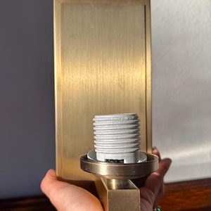 May include: A brass-toned wall sconce with a rectangular backplate and a cylindrical light socket. The sconce has a brushed finish and a simple, modern design. The light socket is white.
