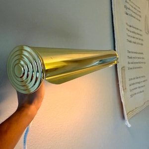 May include: A gold-colored wall-mounted light fixture with a cylindrical design. The fixture has a circular, ridged end and a tapered, elongated body. A white and blue electrical cord is visible.
