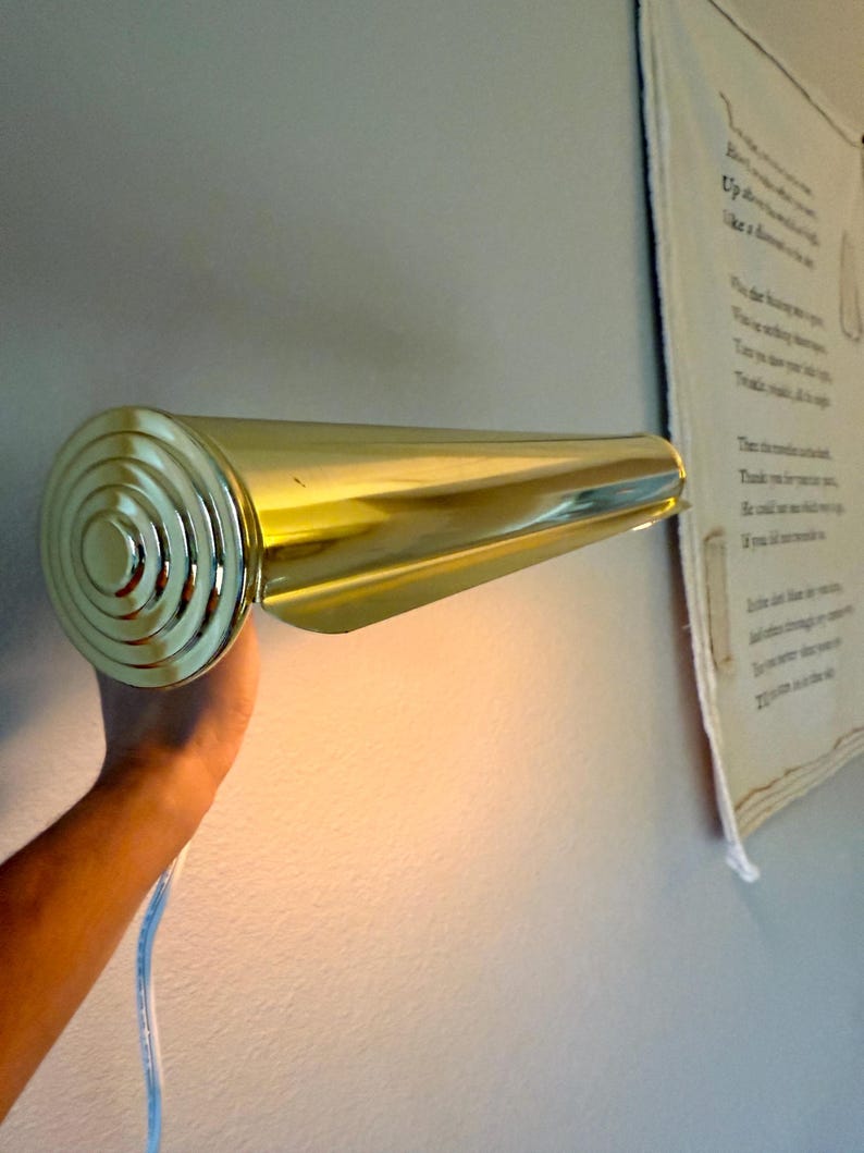 May include: A gold-colored wall-mounted light fixture with a cylindrical design. The fixture has a circular, ridged end and a tapered, elongated body. A white and blue electrical cord is visible.