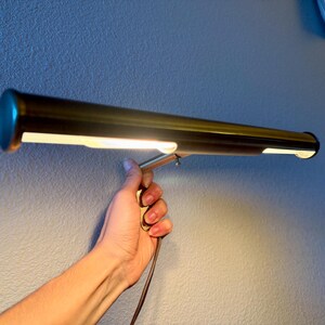 May include: A brass-colored, rectangular lamp with a cylindrical body and a bright, white light. The lamp is held by a hand against a textured, light blue wall. The lamp has a small, round, teal-colored end cap.