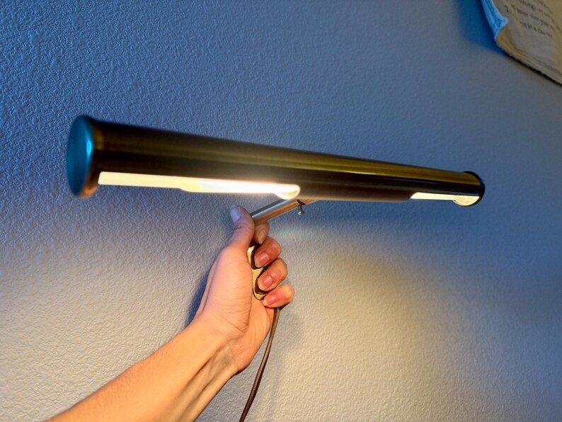 May include: A brass-colored, rectangular lamp with a cylindrical body and a bright, white light. The lamp is held by a hand against a textured, light blue wall. The lamp has a small, round, teal-colored end cap.