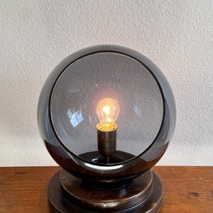 May include: A spherical, dark gray glass table lamp with a warm, glowing lightbulb. The lamp sits on a tiered, dark brown base. The lamp's design is modern and minimalist, suitable for a living room or bedroom.