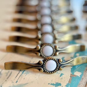 May include: A series of drawer pulls arranged in a line, each featuring an antique brass finish with a curved metal handle and a central circular white ceramic inlay surrounded by a decorative beaded border detail on the metal frame.