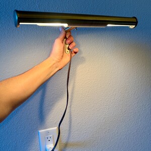 May include: A brass-toned picture light with a rectangular design, held against a blue wall. The light is illuminated, and a brown power cord is connected to a wall outlet. The light fixture is designed to illuminate artwork.