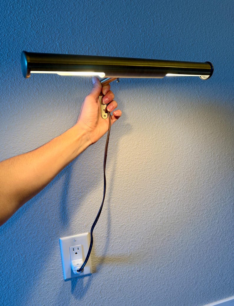 May include: A brass-toned picture light with a rectangular design, held against a blue wall. The light is illuminated, and a brown power cord is connected to a wall outlet. The light fixture is designed to illuminate artwork.