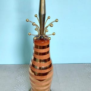 K&ouml;nnte beinhalten: Vintage Tischlampe mit Messing- und Holzdesign. Die Lampe hat einen zylindrischen Sockel mit abwechselnden horizontalen Streifen aus Holz und Messing. Die Oberseite hat eine Messing-Finial und dekorative Arme mit kleinen Messingkugeln.