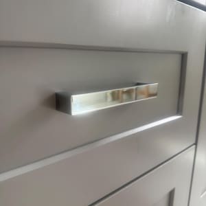 May include: A rectangular, polished chrome cabinet handle with a reflective, mirror-like finish attached to a flat, matte gray drawer front. The handle features clean, sharp edges and a horizontal orientation, contrasting with the smooth gray surface.
